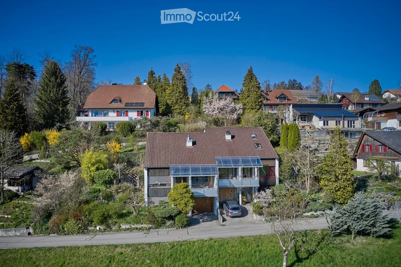 A modern two-story house with a red-tiled roof, large windows, and a glass extension. The house is surrounded by greenery and has a driveway with a parked car and a garage.