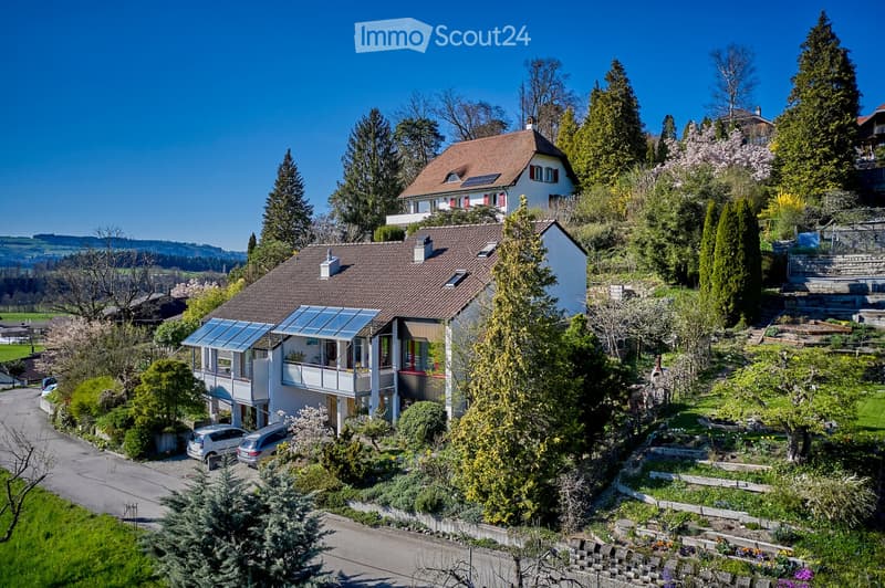 A two-story house with a tiled roof, solar panels, and a well-maintained garden with trees and shrubs. The house features a balcony and a driveway with parked cars.