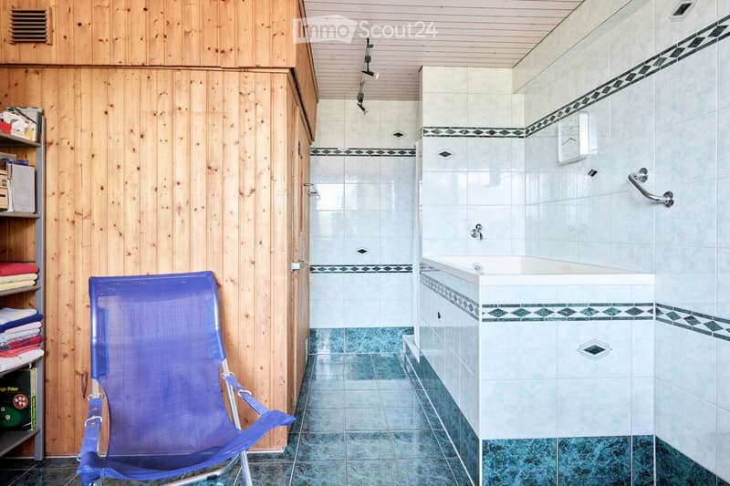 Bathroom with a bathtub, tiled walls, a blue folding chair, and wooden shelves holding towels and books.