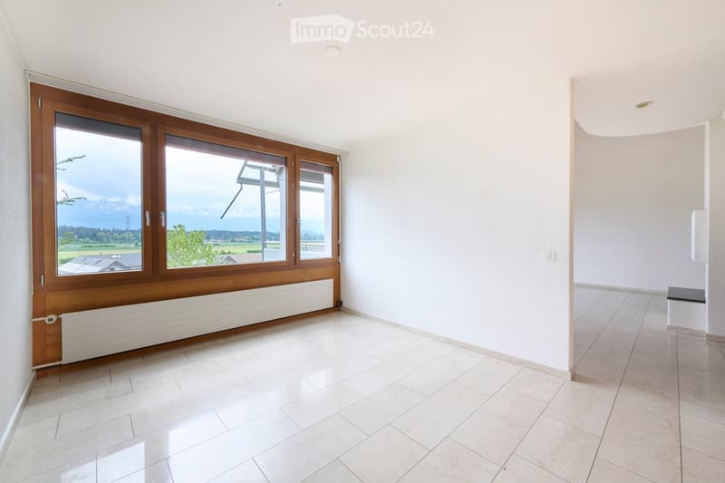 Empty room with white walls, tiled floor, large wooden-framed windows offering a scenic view, and a radiator below the windows.