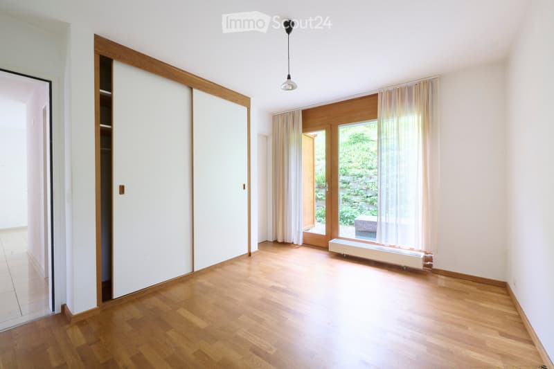 Empty room with wooden flooring, white walls, large window with curtains, sliding glass door leading to outdoor area, and built-in closet with wooden trim.