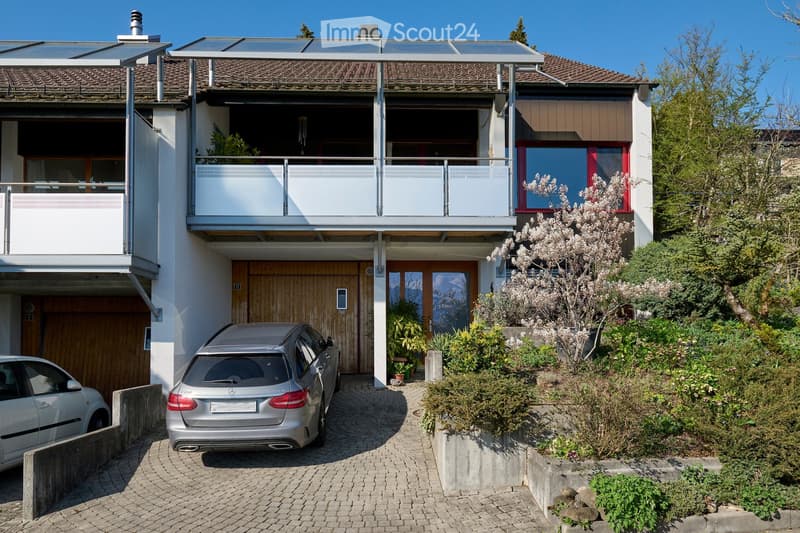 Modern two-story house with a balcony, garage, and solar panels on the roof. The house features large windows, a wooden garage door, and is surrounded by greenery and a paved driveway with parked cars.
