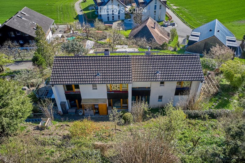A modern two-story house with a tiled roof, large windows, and a raised ground floor, surrounded by greenery and neighboring houses.