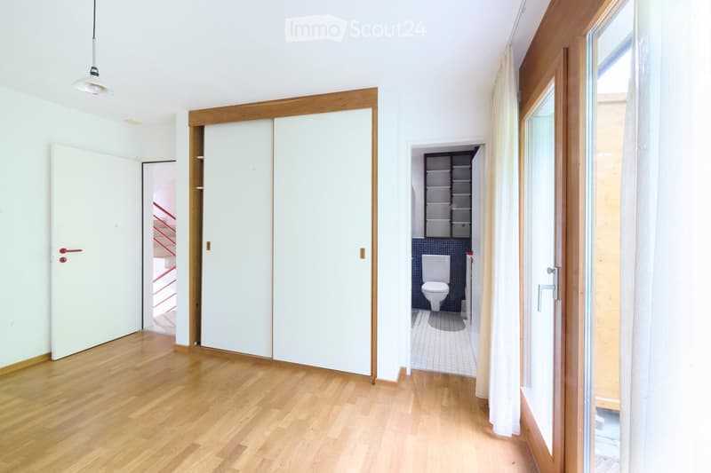 Bright hallway with wooden flooring, white walls, and large sliding doors leading to a bathroom and another room. The bathroom has a toilet, tiled walls, and a small shelf unit.