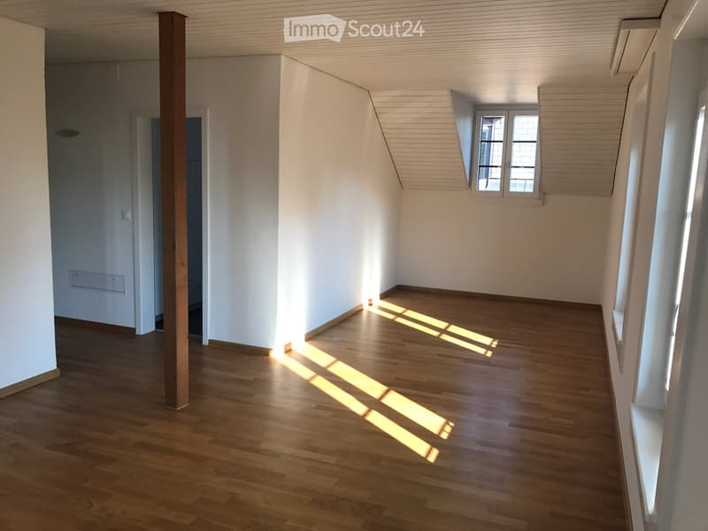 Empty room with wooden flooring, white walls, and large windows allowing natural light to enter. The room features a wooden pillar and a door leading to another space.