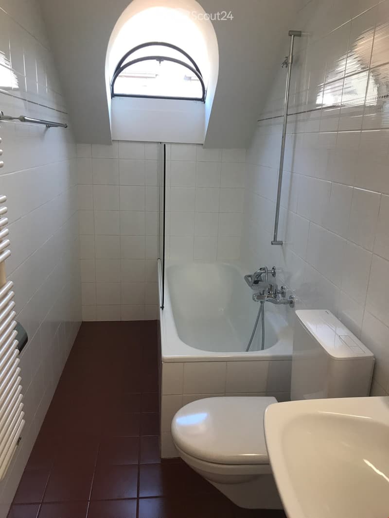 Modern bathroom with white tiles, a bathtub, toilet, sink, and a radiator. Features a large window with blinds and a showerhead above the bathtub.