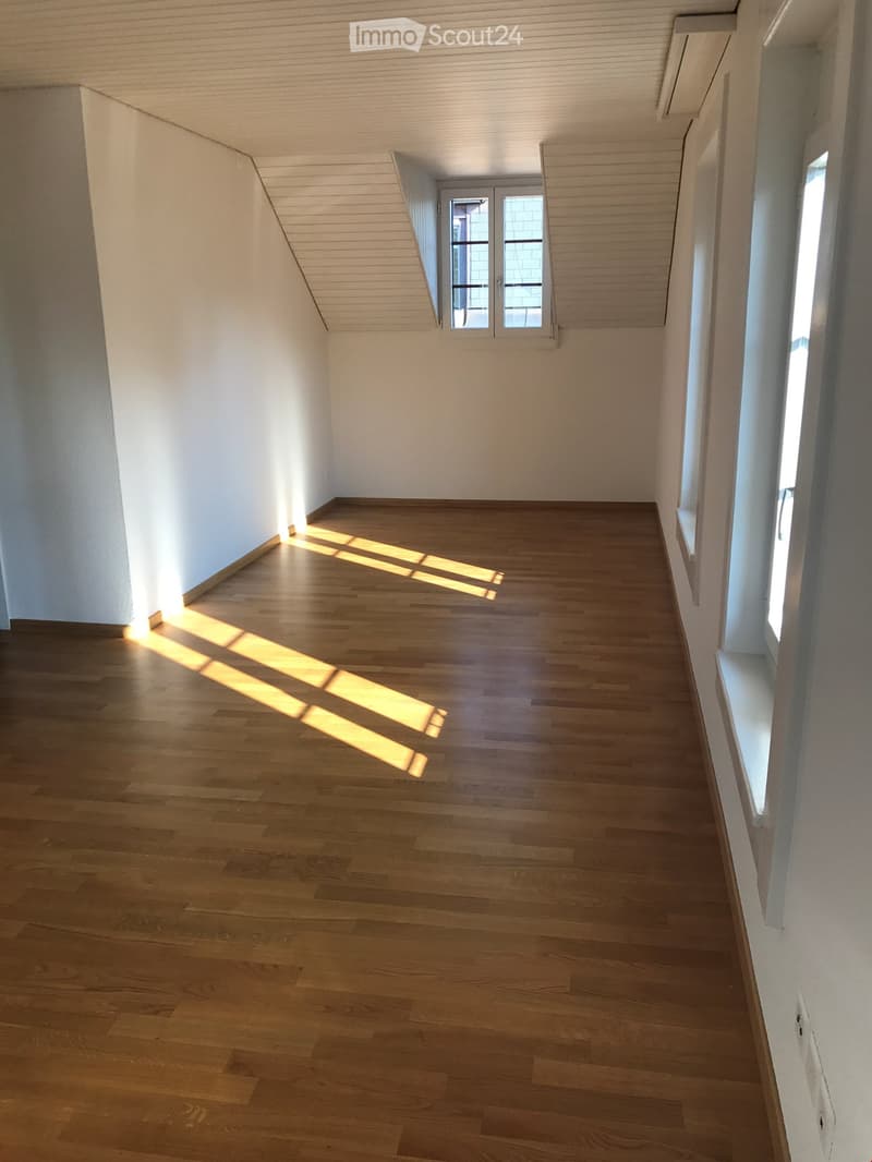Empty room with wooden flooring and white walls, featuring natural light from windows and a door leading to another space.