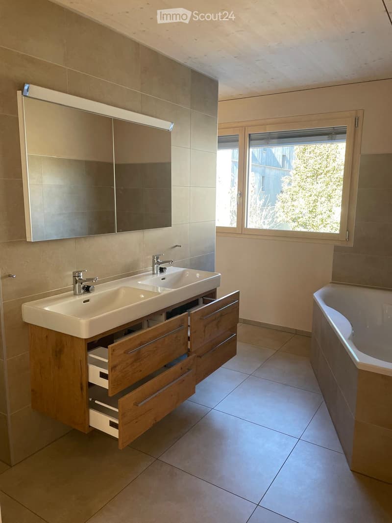 Modern bathroom with a double sink, wooden cabinet, mirror, and bathtub, featuring tiled walls and a large window.