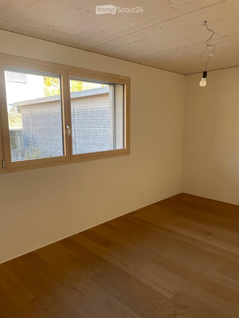 Empty room with wooden flooring, light-colored walls, and large windows with blinds. A single light fixture hangs from the ceiling.