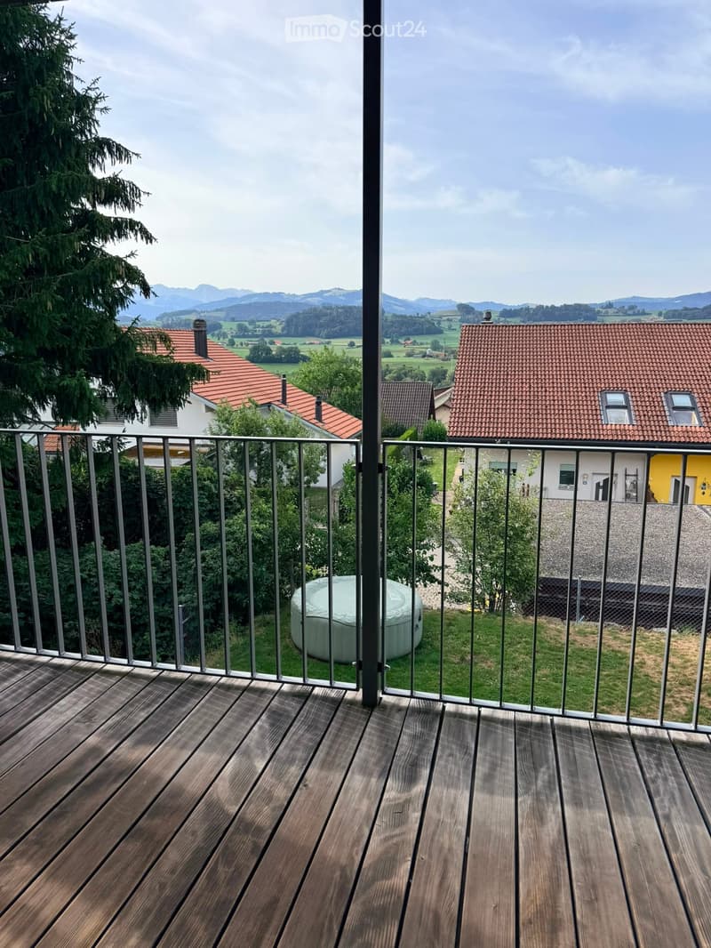 Wooden deck with metal railing, overlooking a scenic countryside with houses, trees, and mountains.