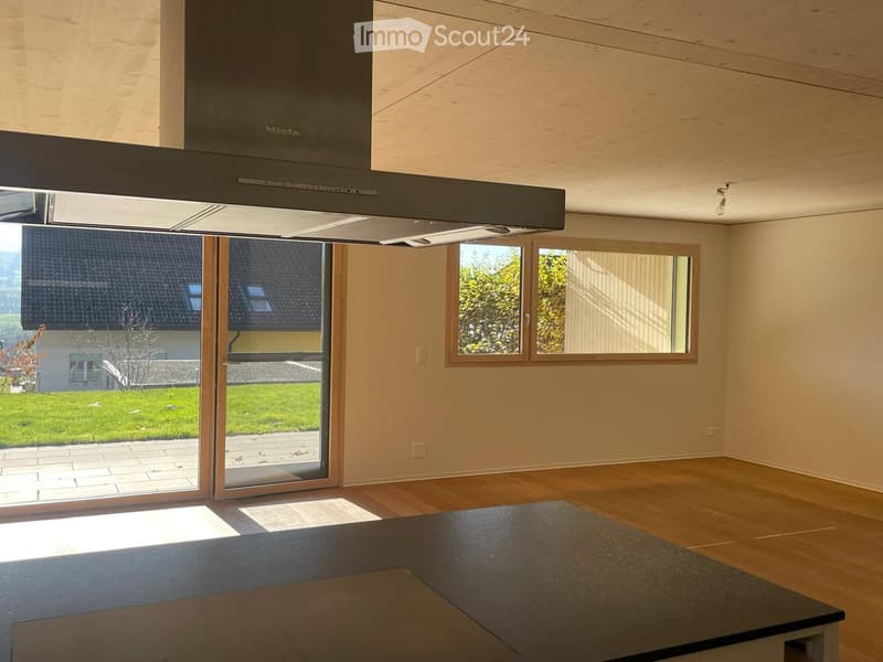 Empty kitchen with wooden flooring, large windows, and a modern range hood. No furniture or appliances are present.