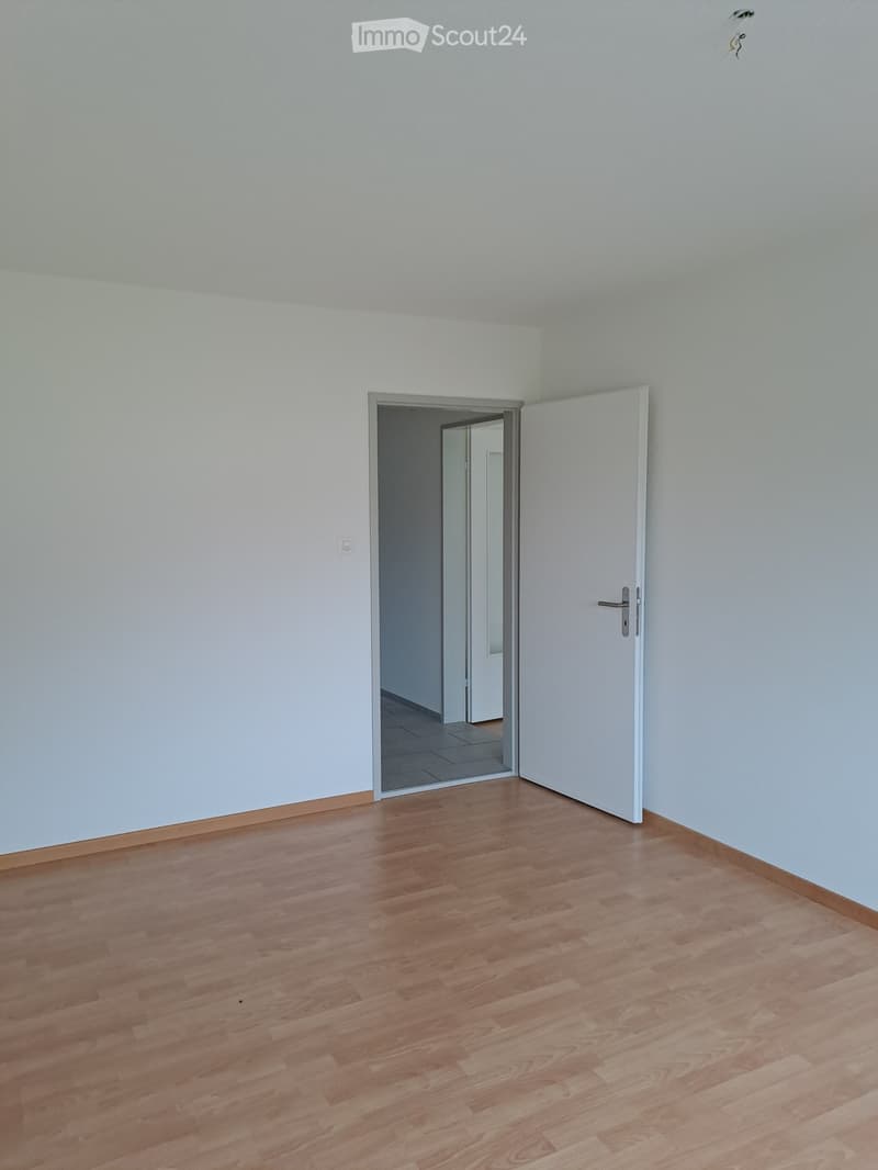 Empty room with wooden floors, white walls, an open door, and a hallway leading to another room.