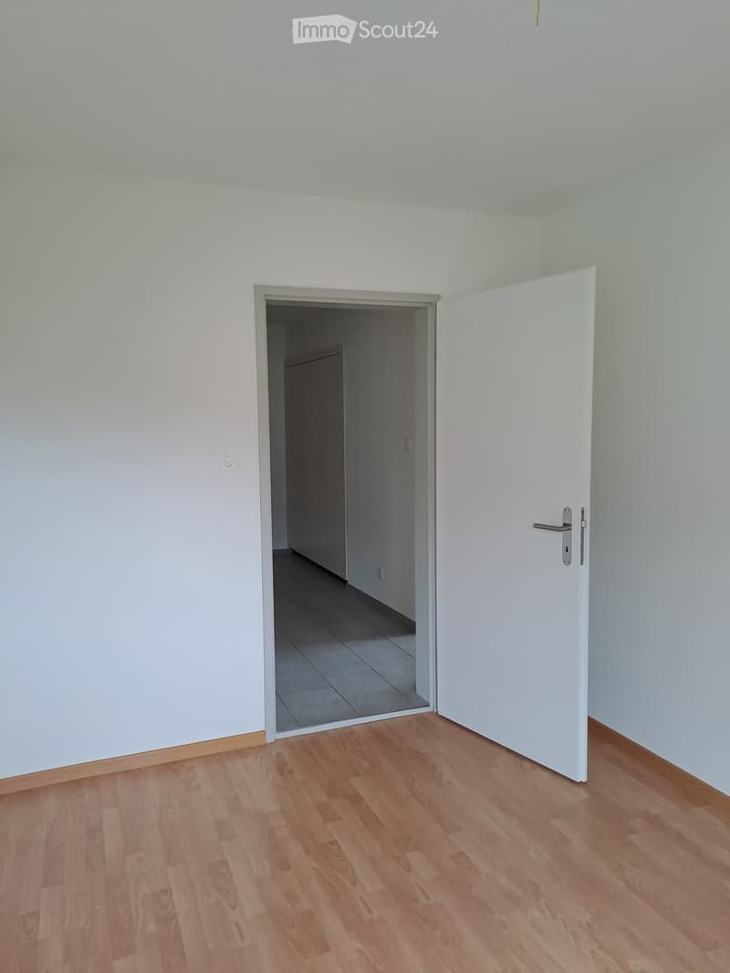 Empty room with wooden floor, white walls, tiled entrance area, open door to another room, power outlet on the wall.