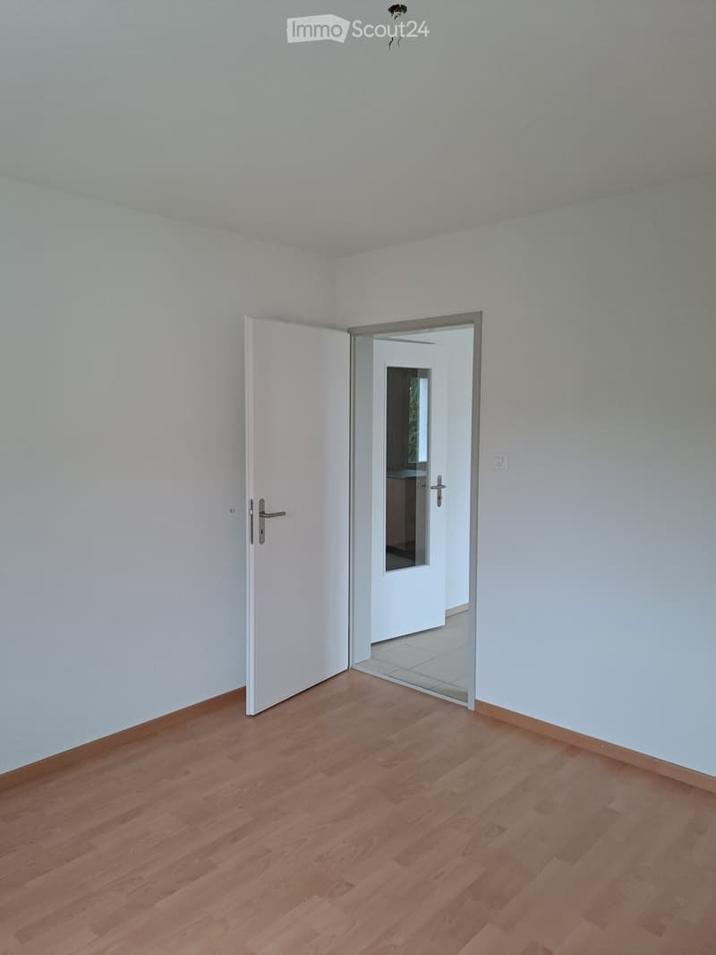 An empty room with white walls, wooden floors, a white door, an open door, and a small hallway.