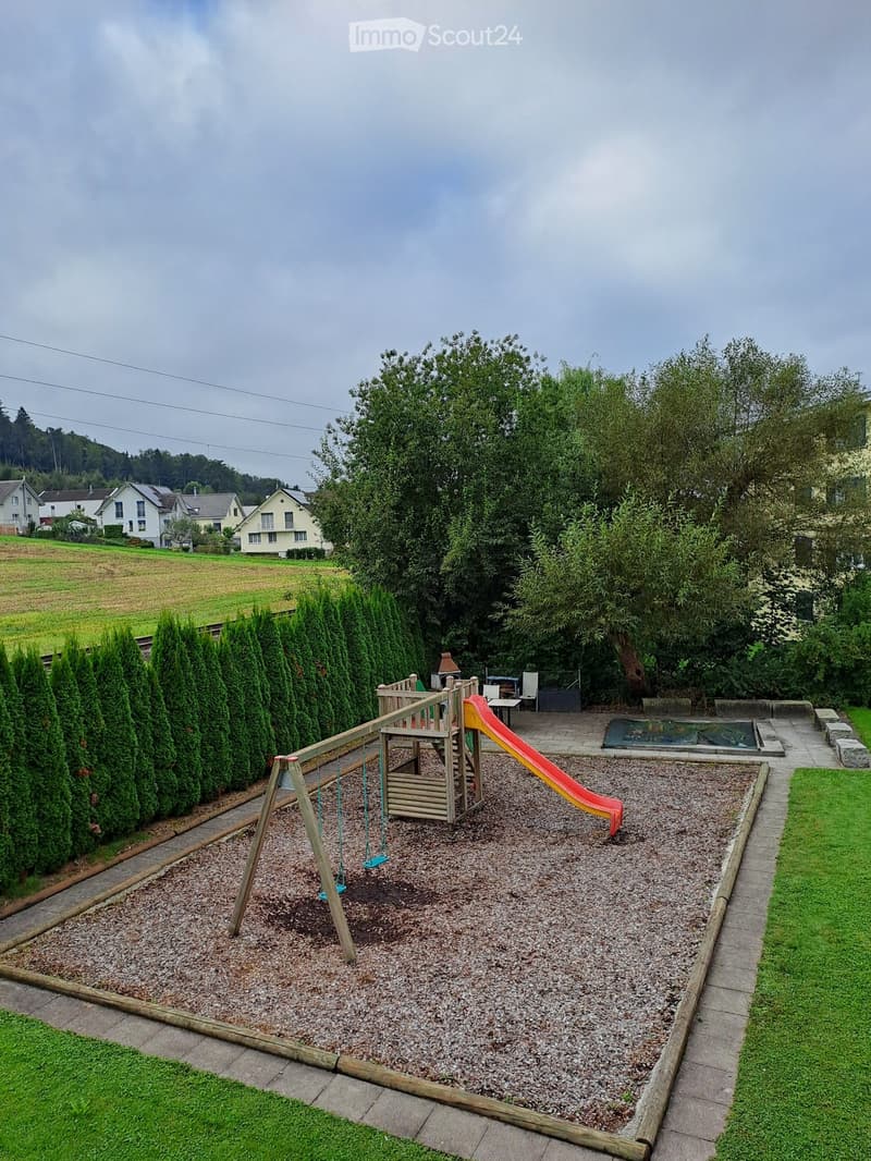 Playground with slide, swings, gravel surface, bordered by hedge, trees, small grass areas, and path