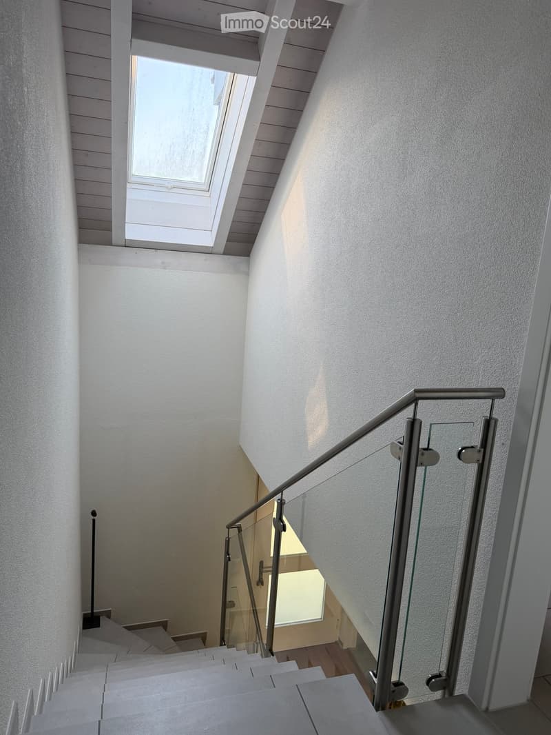 Modern staircase with glass railing and wooden steps, leading to a bright room with large windows.