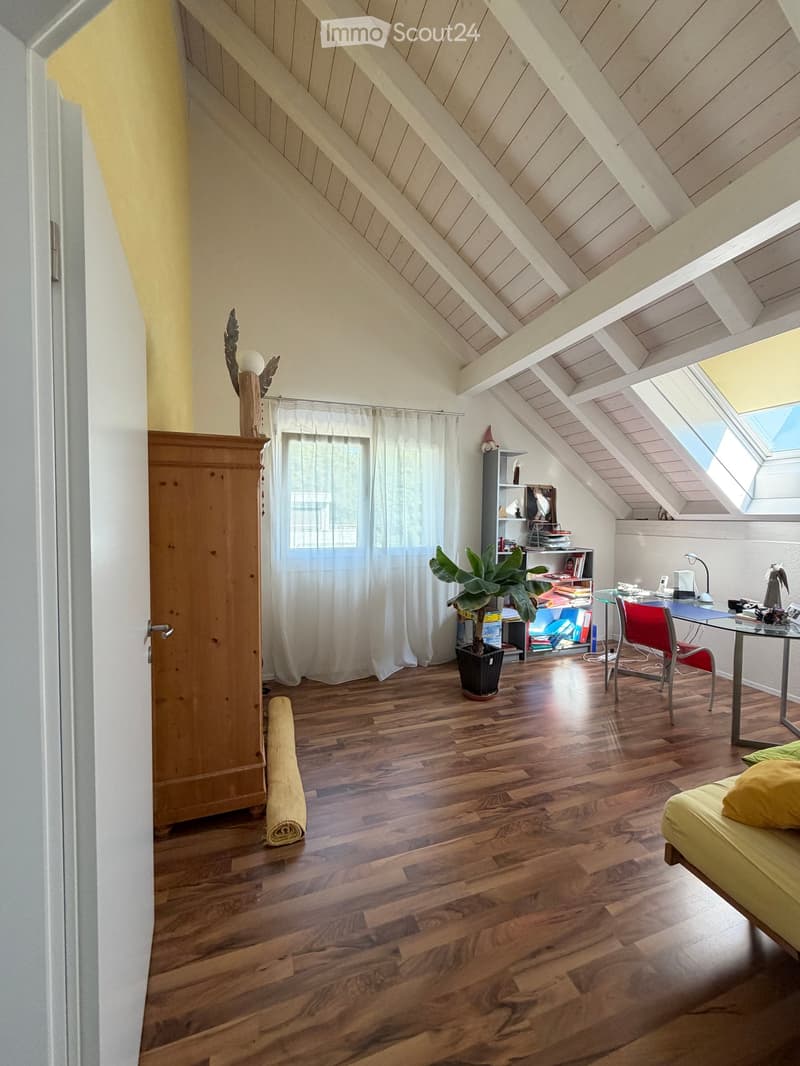 Bright attic room with wooden floors, slanted wooden ceiling, large windows, and skylight. Contains a yellow sofa, glass table, red chair, potted plant, wooden cabinet, and bookshelf.