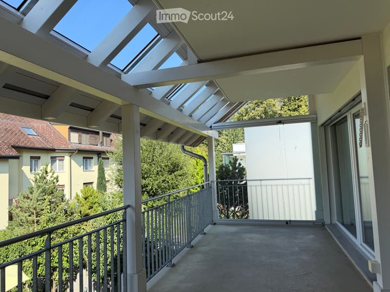 Balcony with a modern glass roof structure, white railings, and a view of surrounding greenery and neighboring buildings.