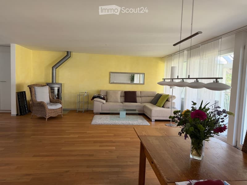 Bright living room with wooden flooring, a beige sectional sofa, a wicker armchair, a glass coffee table, a large flower vase on a wooden side table, and a modern floor lamp.