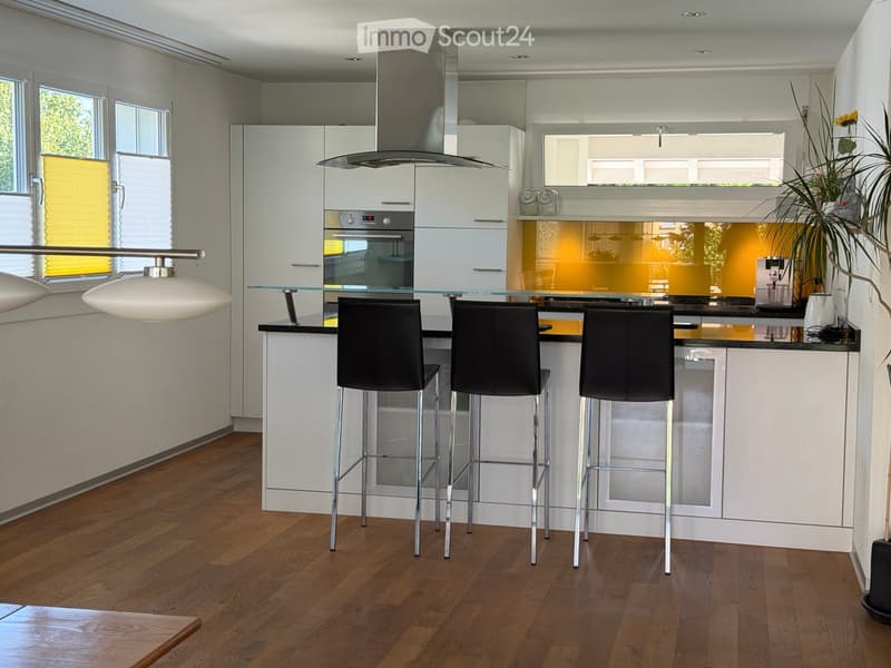 Modern kitchen with white cabinets, glass island, three black bar stools, pendant light, and a potted plant on the counter.
