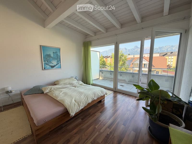 Bright bedroom with wooden flooring, large windows offering a mountain view, a double bed with pillows and blankets, a nightstand with a lamp, and a potted plant near the window.