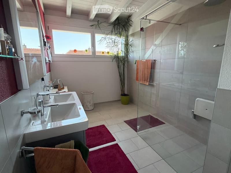 Modern bathroom with double sink, glass-enclosed shower, and large windows offering a view of the roof.