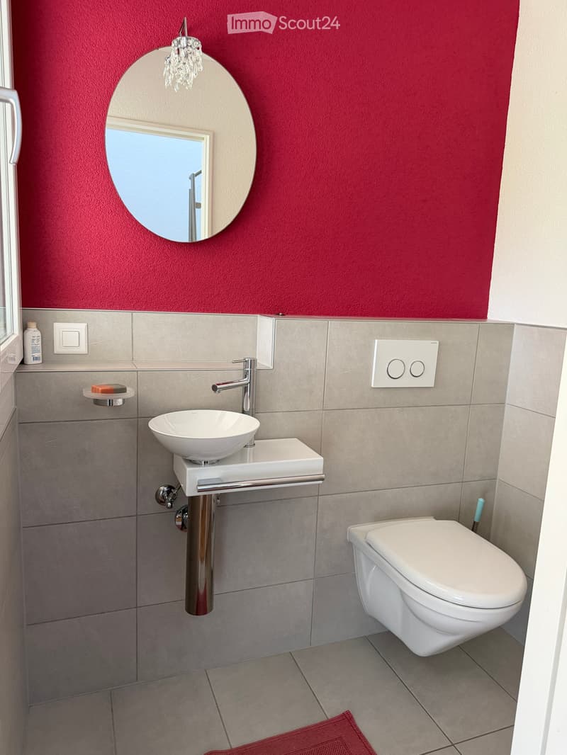 Modern bathroom with a white sink, chrome faucet, toilet, round mirror with a crystal light fixture, and a red bath mat on the floor.