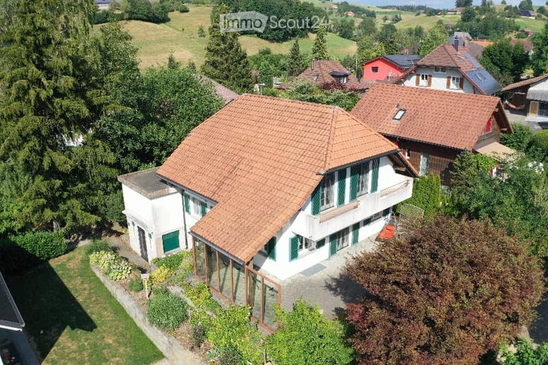 Aerial view of a two-story house with a red-tiled roof, white walls, green shutters, and a balcony. The house is surrounded by a well-maintained garden and trees, with neighboring houses visible in the background.