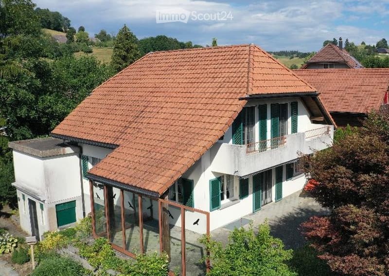 A two-story house with a red-tiled roof, white walls, green shutters, and a balcony. The house is surrounded by trees and greenery, with a driveway leading to the entrance.