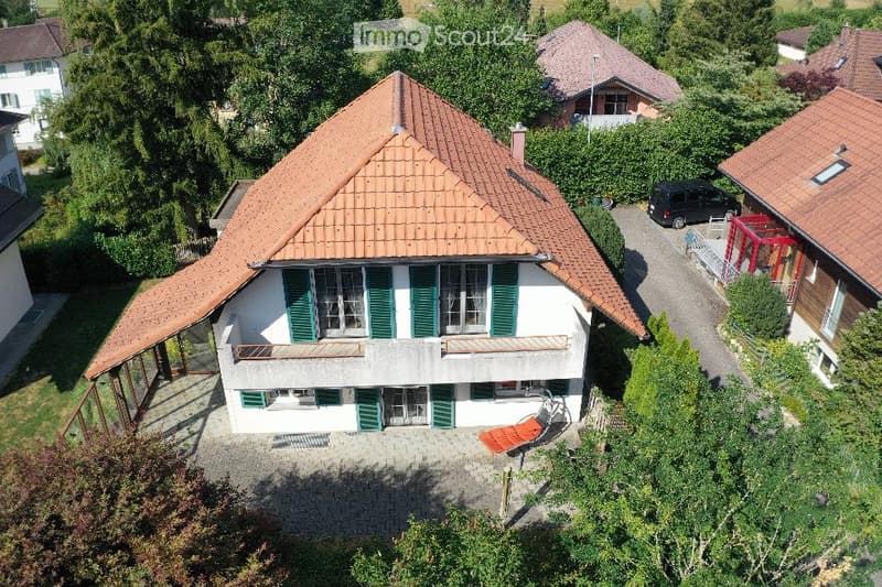 Aerial view of a two-story house with a red-tiled roof, white walls, green shutters, and multiple windows. The house has a covered patio area with outdoor furniture and is surrounded by greenery and neighboring houses.