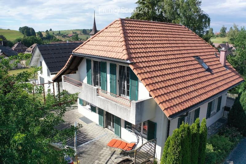 A two-story house with a red-tiled roof, white walls, green shutters, and a balcony overlooking a scenic countryside view.