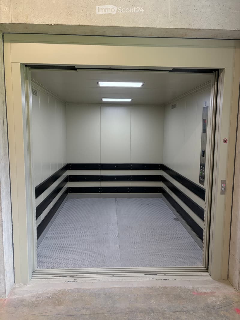 Elevator shaft with white walls, black trim, and a textured floor, viewed from the open elevator door.