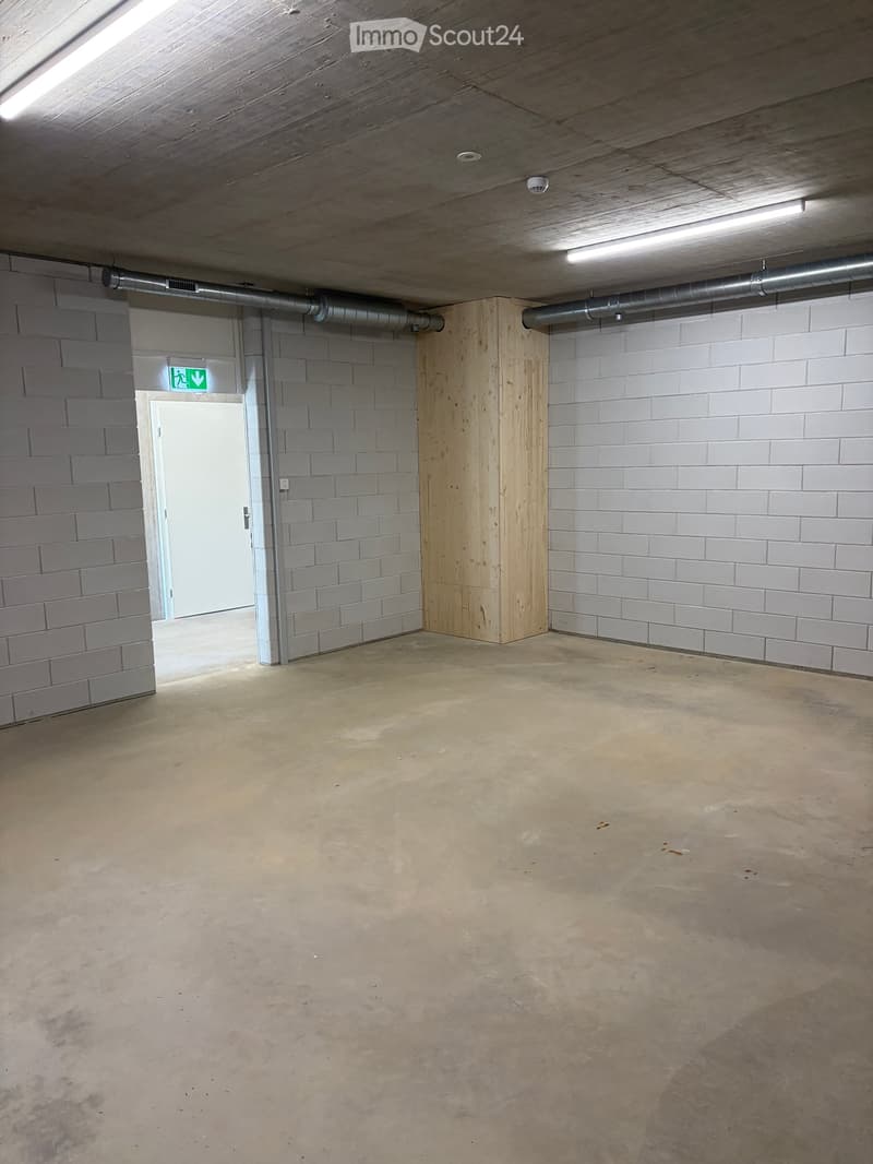 Empty room with concrete floor, grey brick walls, wooden paneling, and a white door with an exit sign above it.