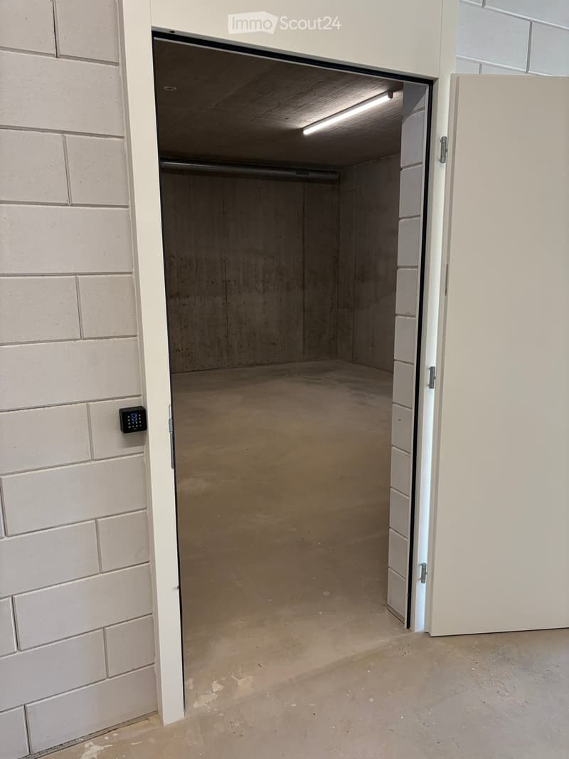 Empty garage with concrete floor, white brick walls, and an open door leading to a concrete-floored space.