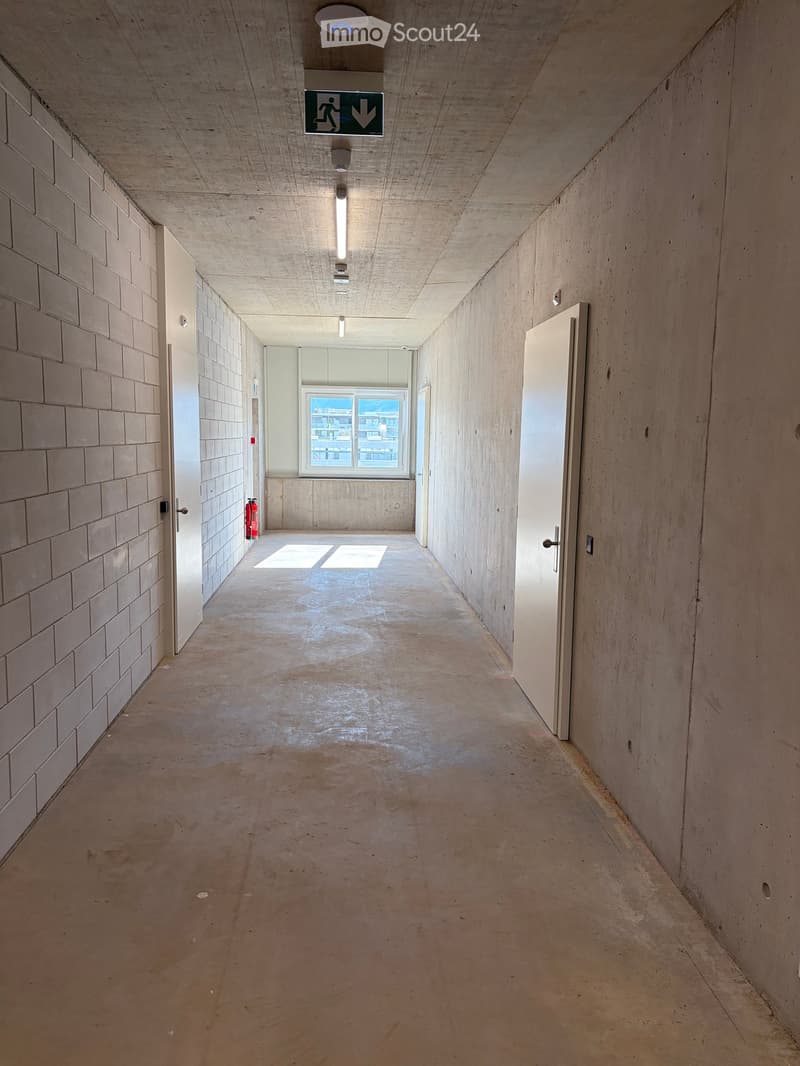 Empty hallway with concrete walls and floor, white doors, a window at the end, and a fire extinguisher on the left side.