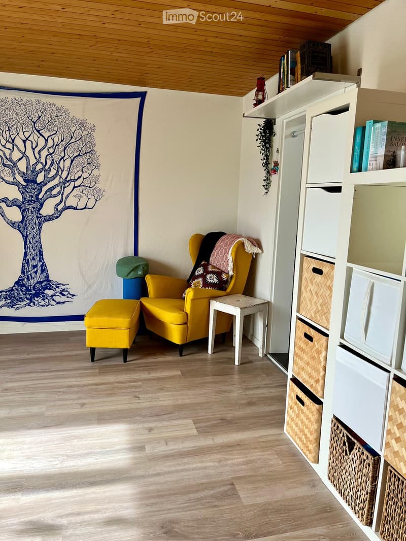 A cozy living room with a yellow armchair, matching yellow stool, small white table, and a white shelving unit with books and decorative items. The room features a large tapestry with a tree design on one wall and a wooden floor.