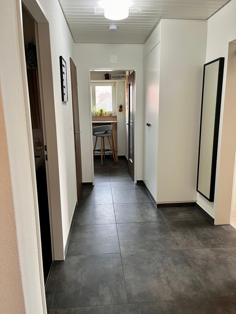 Clean, modern hallway with gray tiled flooring, white walls, and recessed lighting. Features include a small seating area with a wooden table, gray chair, and a potted plant visible through an open doorway.
