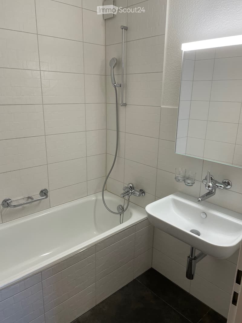 Modern bathroom with a bathtub, sink, mirror, and showerhead. Features white tiled walls and a built-in handrail next to the bathtub.