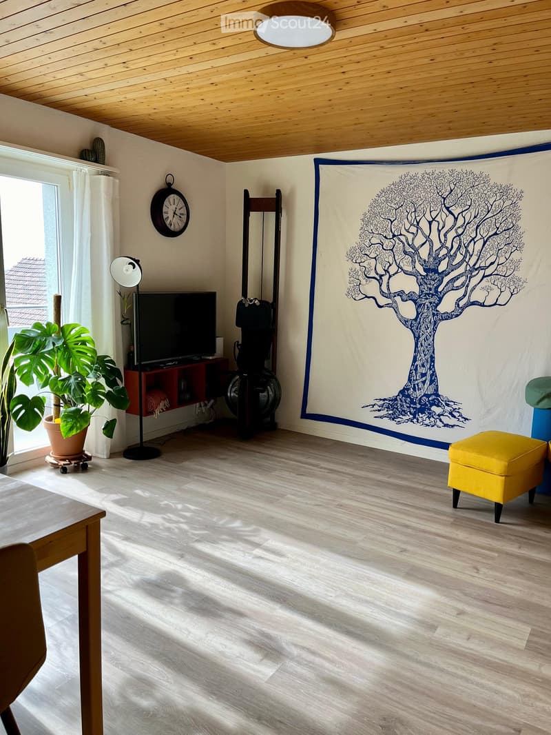 Bright living room with wooden flooring, a large window, a potted plant, a television on a stand, a yellow stool, and a tapestry with a tree design on the wall.