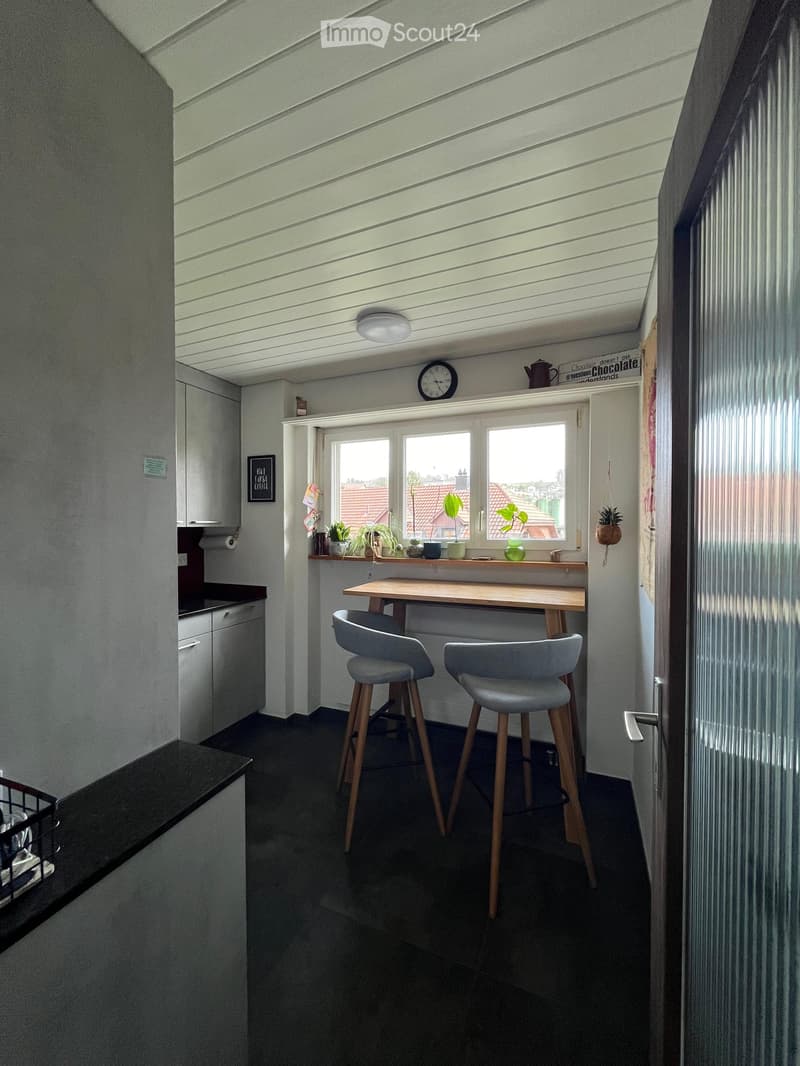 Modern kitchen with a wooden table, two grey stools, white cabinets, and a window with potted plants on the sill.