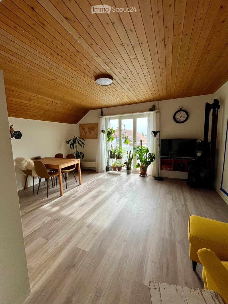 Bright living room with wooden flooring, slanted wooden ceiling, large window with white curtains, sofa, dining table with chairs, television, and potted plants.