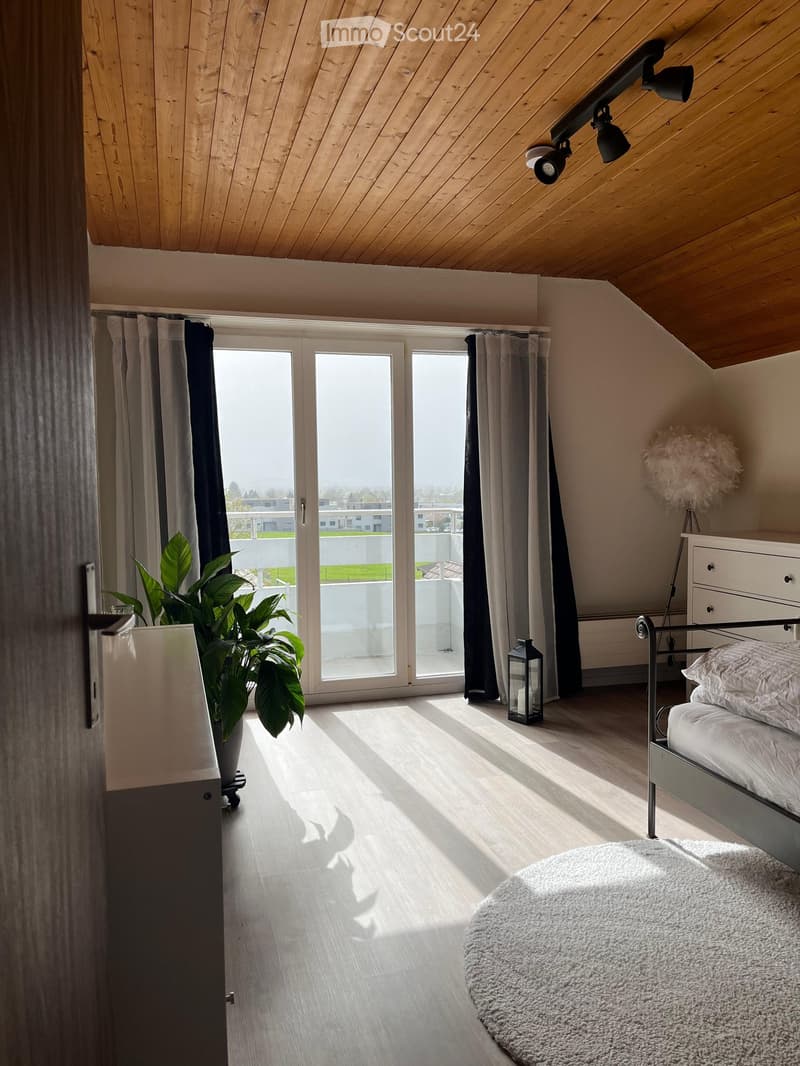 Bright, modern bedroom with wooden floors, large windows, a bed with white bedding, a nightstand, a plant, and a round rug.