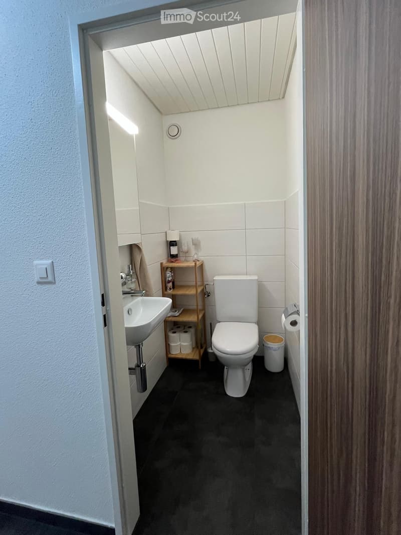 Modern bathroom with white tiles, a toilet, a sink, a wooden shelf holding toilet paper and a bottle, and a small trash bin.