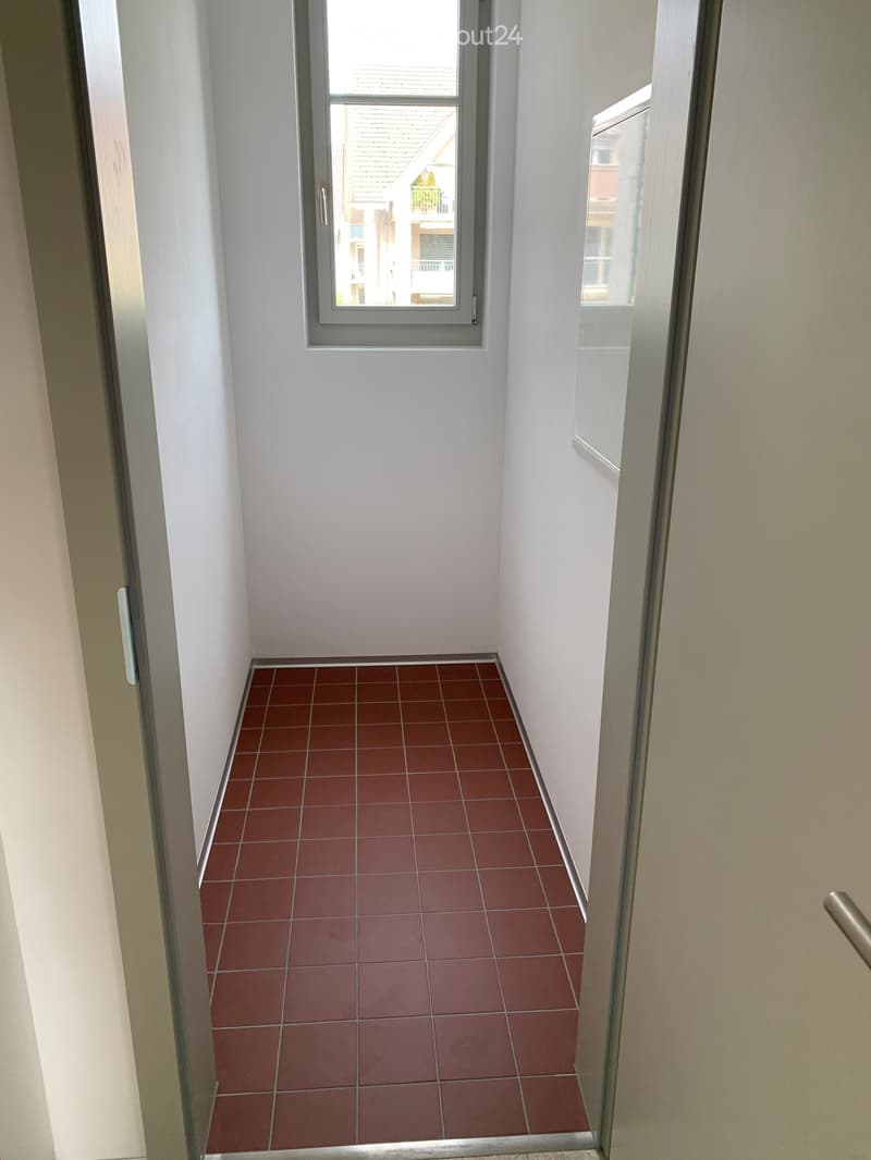Narrow hallway with red tiled floor and white walls, featuring a window at the end showing a view of neighboring buildings.