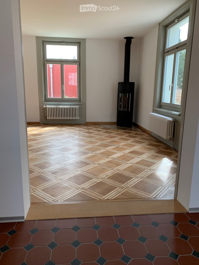 Empty room with wooden floor, fireplace, windows, and radiators.