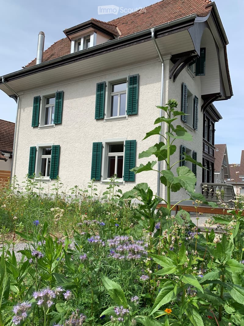 Two-story house with a red-tiled roof, white facade, green shutters, and a garden with flowers and plants in the foreground.