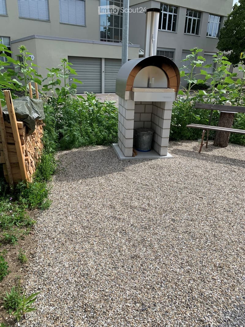 Garden area with a modern outdoor pizza oven, gravel ground, wooden bench, and lush green plants.