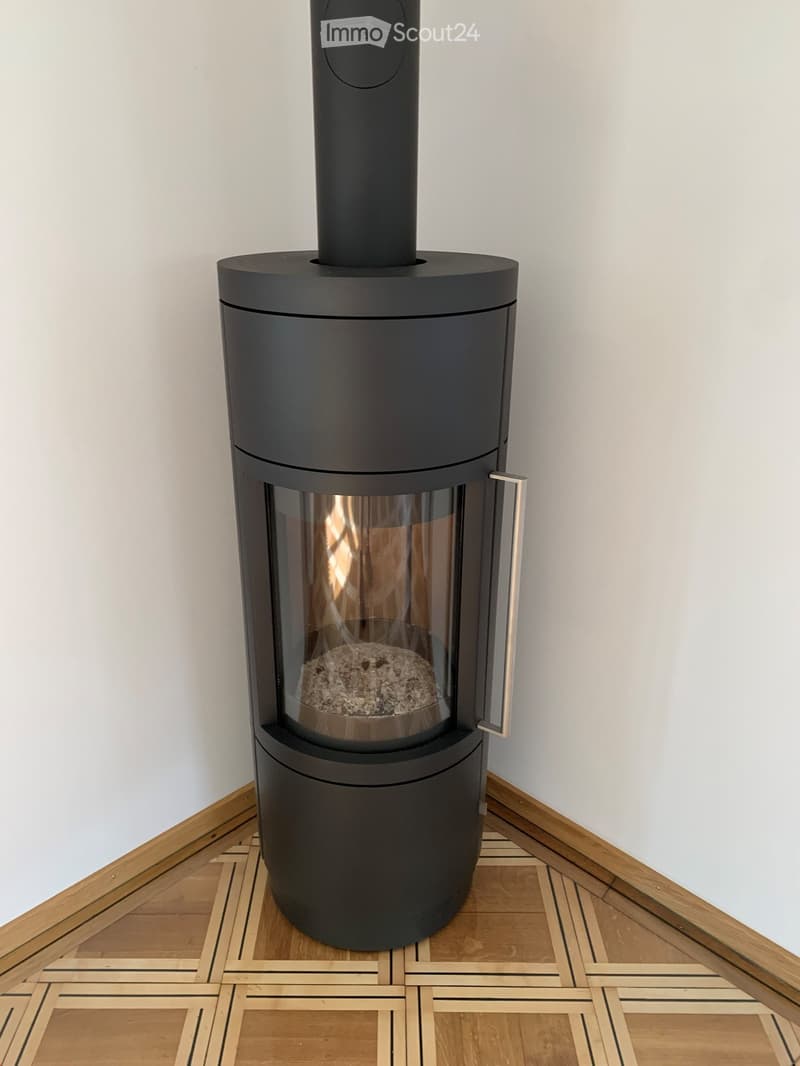 Modern black cylindrical wood-burning stove with glass front, placed on a wooden floor in a room with light-colored walls.