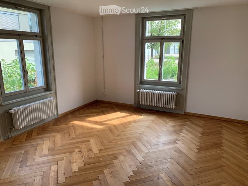 Empty room with wooden herringbone flooring, large windows, and radiators beneath the windows.