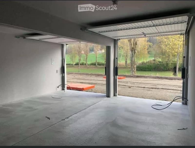Empty garage with concrete floor, large open door leading to outdoor area, visible electrical wiring and a small orange object on the floor.